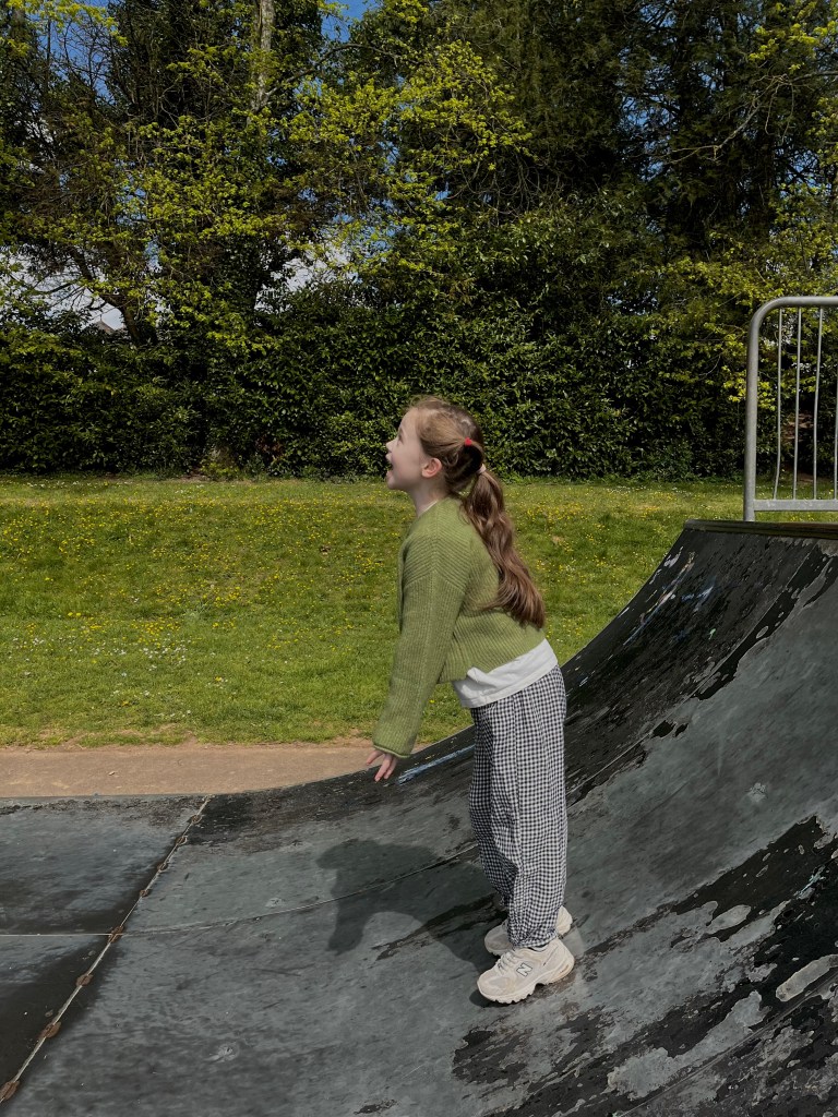A child with long hair wearing a green sweater and checkered pants stands on a skate ramp, looking up with a joyful expression, surrounded by greenery and blue sky.