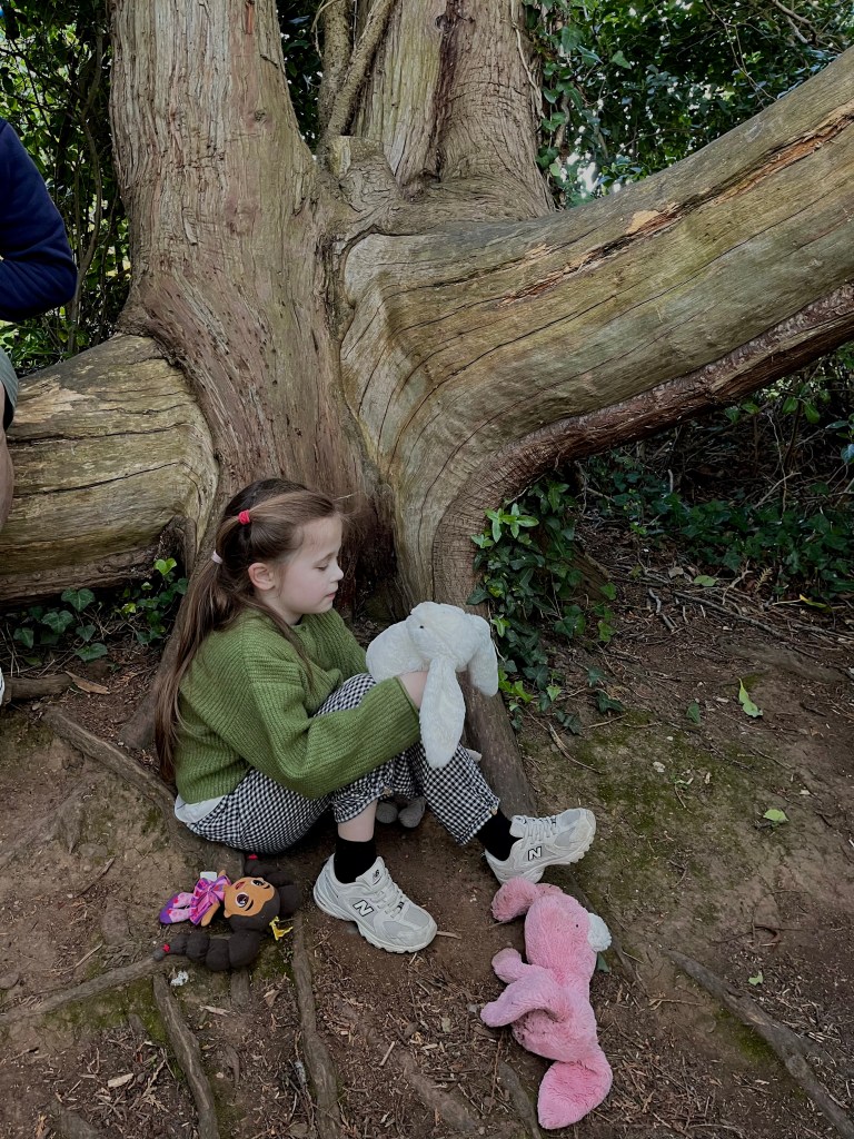 A young girl sitting on the ground next to a large tree, holding a white stuffed bunny. She has long hair and is wearing a green sweater and checkered pants. Various stuffed animals, including a monkey and a pink bunny, are scattered around her.