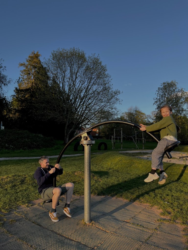 A man and a girl playing on a playground teeter-totter during sunset, surrounded by green grass and trees.