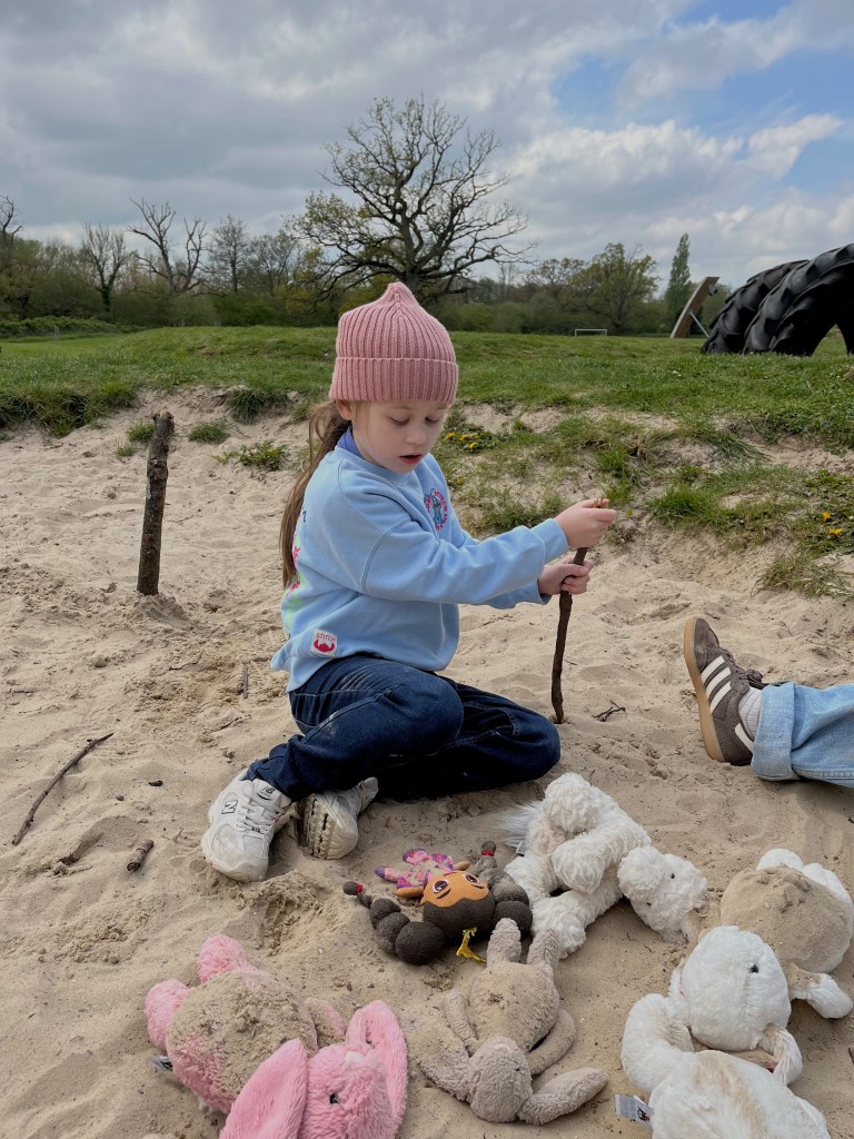 A young child sitting on sandy ground, engaged in play with sticks and several stuffed animals, wearing a pink beanie and a light blue sweatshirt, with trees and a cloudy sky in the background.