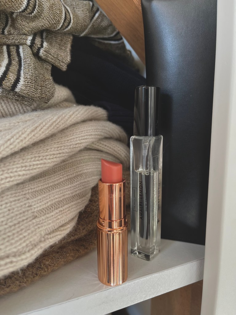 A close-up image of a peach lipstick in a rose gold tube and a clear perfume bottle labeled 'Amalfi Lemon' placed on a shelf with neatly folded knitwear.
