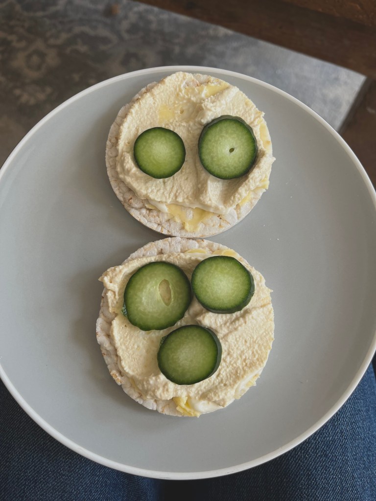 A plate with two rice cakes topped with hummus and slices of cucumber arranged in a playful manner, set against a textured background.