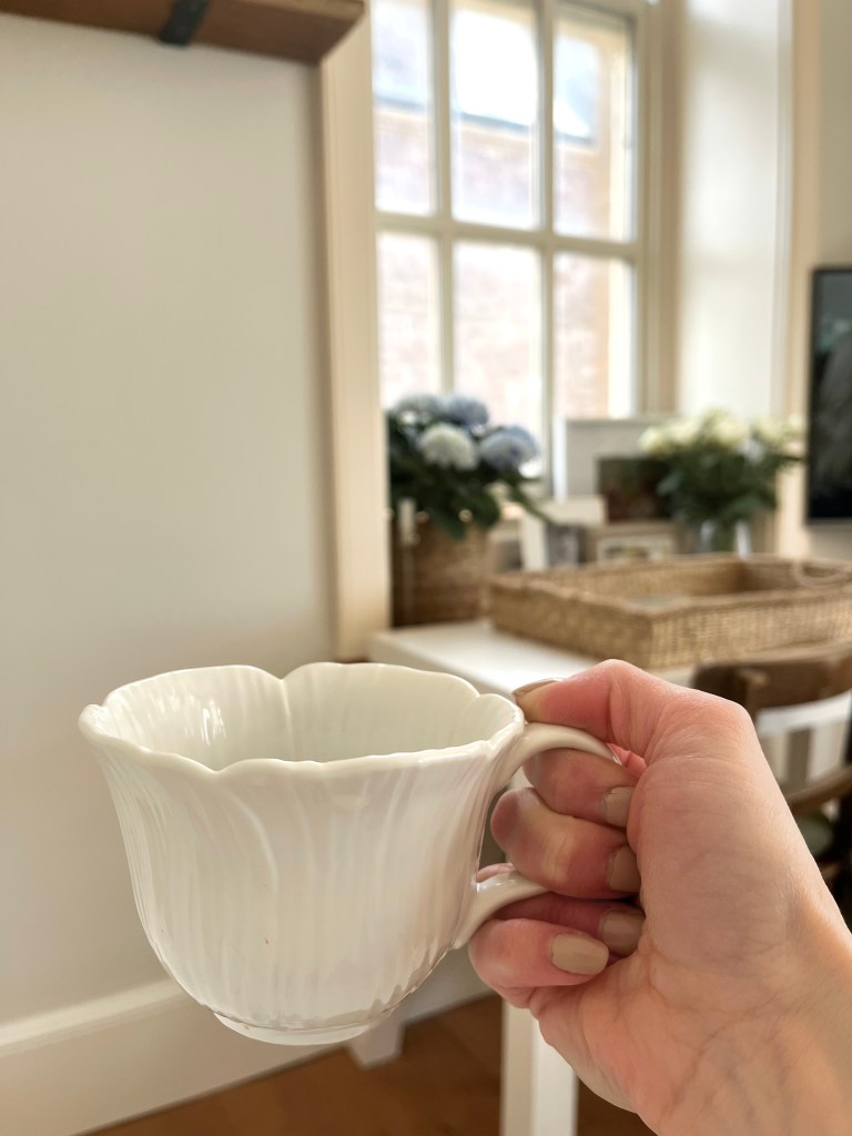 A hand holding a delicate white porcelain cup with a floral design, set against a light-filled room with soft furnishings and a decorative basket.